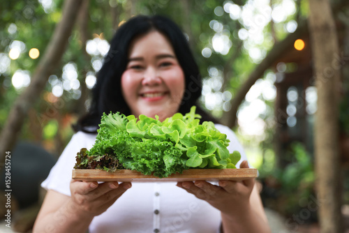 A cheerful plus-size woman holding a wooden tray filled with fresh green lettuce, symbolizing healthy eating and a balanced lifestyle