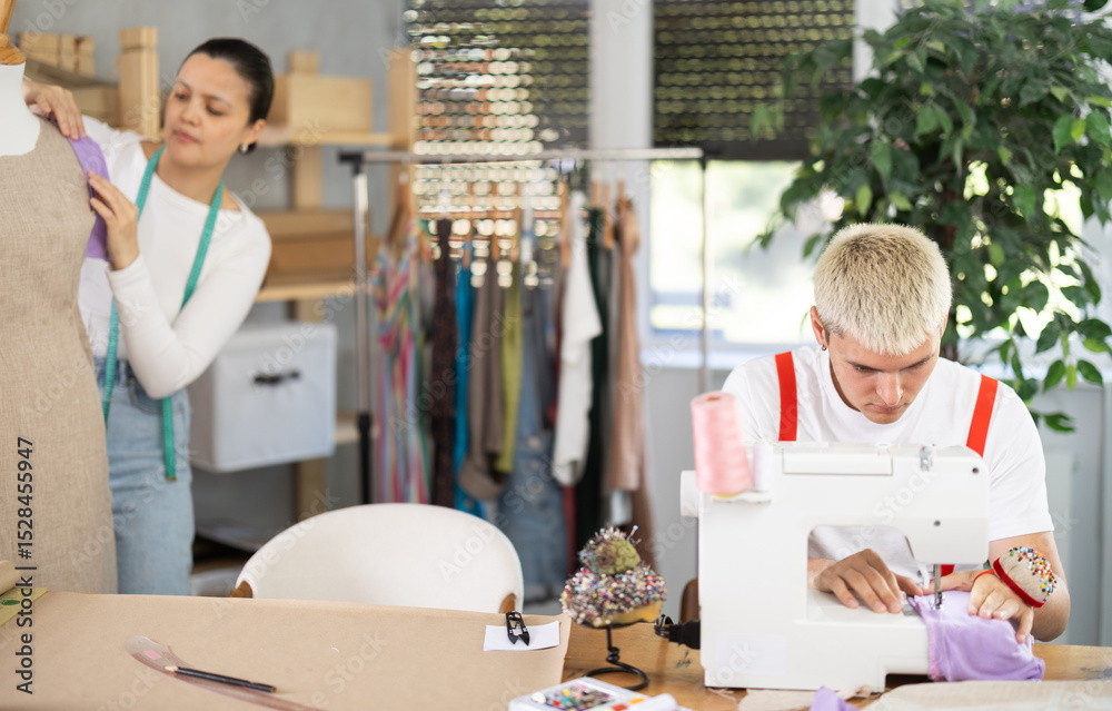Obraz premium Attentive young tailor using sewing-machine while Asian girl working with mannequin in sewing workshop