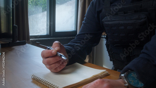 Closeup of a uniformed officer holding a pen and writing in a notebook near a window, possibly documenting an incident or report.