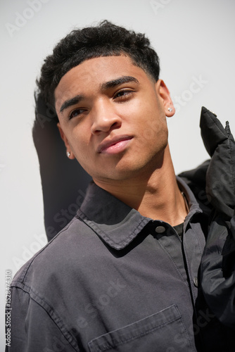 Confident and stylish Dominican man with full lips poses for a portrait, leaning casually against a white wall. His attractive features and self-assured expression reflect youthful charisma and ease.