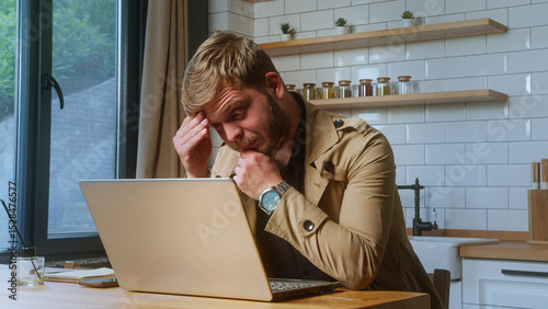 Frustrated man in beige jacket holds his forehead in stress while staring at a laptop, indicating pressure or bad news.