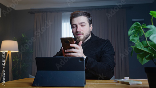 Young man sits in a modern room holding phone in one hand while looking at a tablet on the desk. Focused man reviewing documents or working on administrative tasks