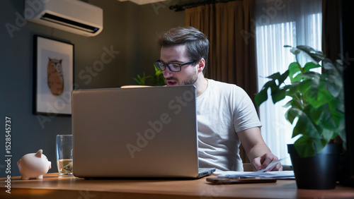 A man wearing glasses participates in an online video call meeting from his home. Seated at a desk and speaking, with a laptop work with papers