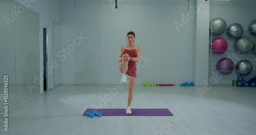 In a bright gym a girl athlete demonstrates a standing knee hug stretch balancing on one leg while holding the other knee to her chest. This exercise promotes flexibility and strength.