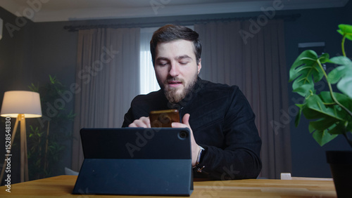 A man sits in a modern room holding phone in one hand while looking at a tablet on the desk. Focused man reviewing documents or working on administrative tasks