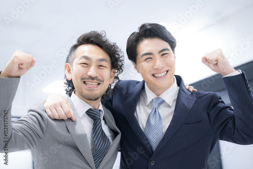 肩を組むガッツポーズをするスーツを着たビジネスマン2人のカメラ目線　成功など楽しい仲良しイメージ