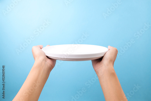Female Hand Presenting Empty White Plate Against Blue Background