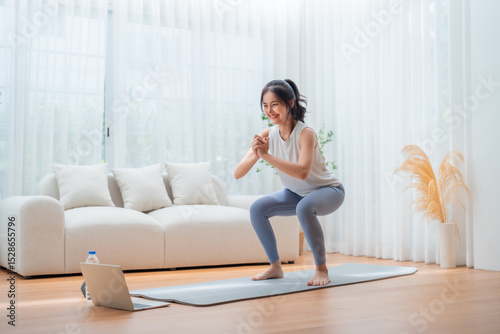 Young Asian beautiful woman doing aerobic exercise at home. Attractive girl doing  workout by squatting, follow instructions video from online trainer for health in house
