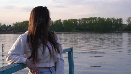 A beautiful girl with long hair stands on a river pier and looks into the distance