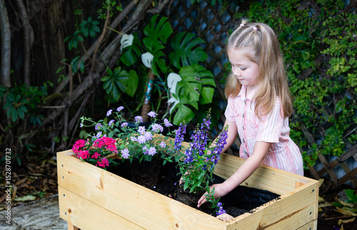 Happy child planting flower pot in backyard – little girl gardening in raised planter box, smiling in front of wooden trellis and green vines, nature education and spring lifestyle concept. 