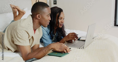 African American couple on bed booking trip online pointing passport at laptop and entering card