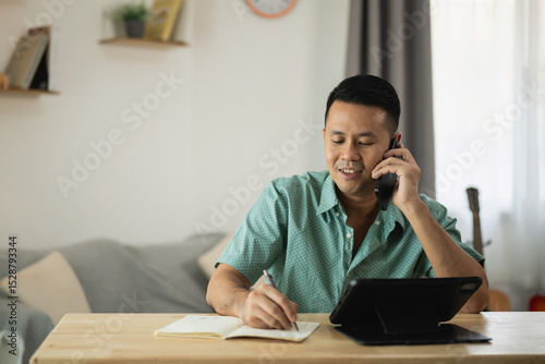Professional man engaged in phone conversation while taking notes at home office desk, showcasing modern workspace and productivity concept in casual attire