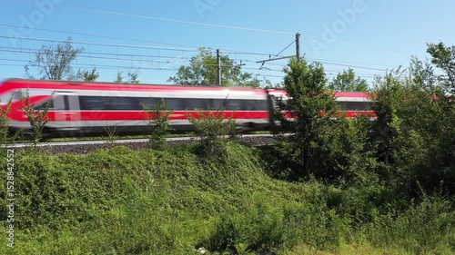 The high-speed train crosses the Italian countryside, its sleek red silhouette standing out against the green trees and bushes.