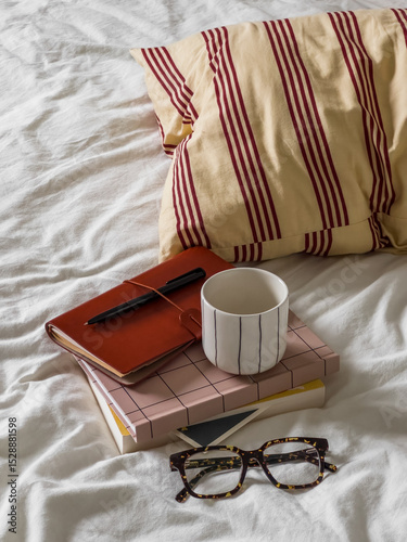 A stack of books, a notebook, a glass of coffee, glasses on a bed with pillows