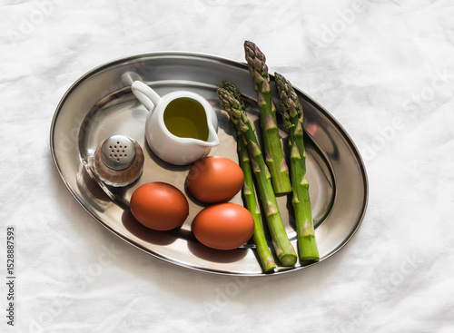 Breakfast ingredients - fresh asparagus, eggs, olive oil, paper on a metal plate on a light background, top view