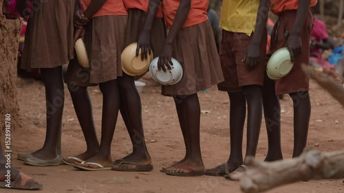 African children stand in line with bowls in their hands to get portion of food during break between classes. Slow motion. Close-up of schoolchildren's feet in flip-flops