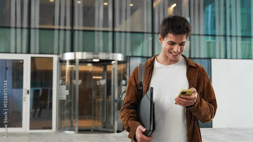 Smiling student standing on university using mobile phone app for exam results, happy looking at the camera Smiling student standing on university using mobile phone app for exam results, happy looking at the camera