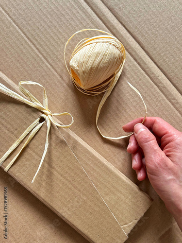 Hand tying natural raffia ribbon around a cardboard package during eco-friendly gift wrapping