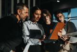 A diverse group of business people are gathered around a digital tablet, collaborating on a project in a modern office setting.