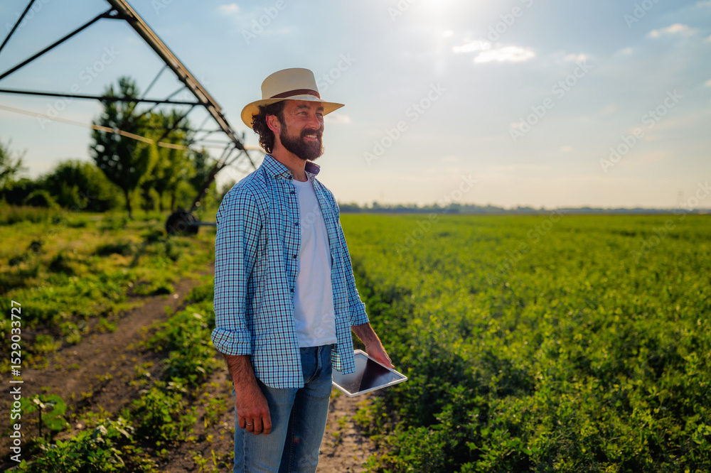 Fototapeta premium Farmer with a straw hat and a beard is holding a tablet and looking at his crops in a cultivated field, with an irrigation system in the background, on a sunny day