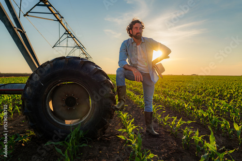 Confident farmer leaning against a tractor wheel, holding his hat while watching over a corn field at sunset, feeling proud of his hard work in agriculture and cultivation