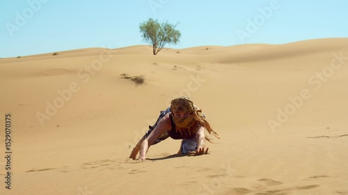 A person in a scarf crawls across scorching sand dunes, enduring heat and fatigue. A lone tree stands in the background under the blazing sky.