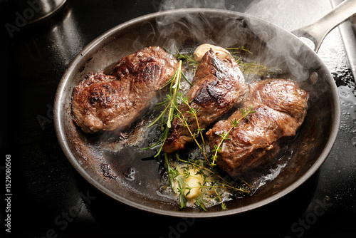 Fresh steaming beef steaks are seared in a carbon steel frying pan with butter, garlic, thyme and rosemary, cooking tasty meat, selected focus
