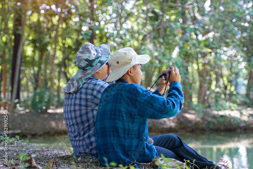 Two Asian boys use binoculars and a camera to study the local birds in a community forest. Soft and selective focus.