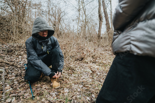 A person dressed in warm outdoor attire bends down to tie their shoelaces in a wooded area marked with fallen leaves, depicting preparation for a journey in nature during a chilly day.