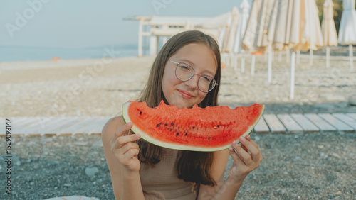 Wallpaper Mural Smiling girl with glasses enjoying a slice of watermelon on a sunny beach day, sitting on pebbles near sun loungers with umbrellas in the background. Torontodigital.ca