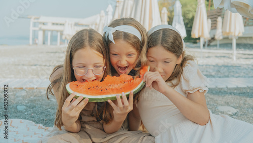 Wallpaper Mural Three happy girls share a big slice of watermelon on a sunny beach day, laughing and enjoying the summer together by the sea with umbrellas in the background. Torontodigital.ca