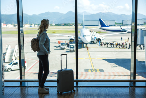 Woman with suitcase waiting at airport terminal before boarding airplane, solo travel passenger ready for flying to summer vacation