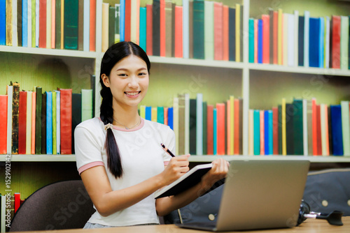 An Asian female university student sits in the library with her laptop, happily researching and working on academic tasks in a calm and focused environment.