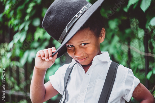 boy dressed in the style of the 20s lifts his hat, holding it by the brim, looking at the camera from under his brows