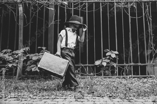 A boy dressed in the style of the 20s dances on the street holding the brim of his hat with a suitcase in his hand