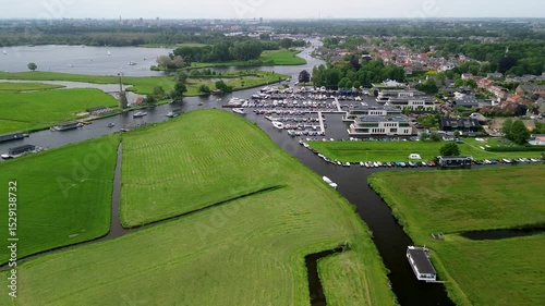 A narrow water channel winds through a green forest, reflecting the surrounding trees. A beautiful Dutch village in the background. A large pier with yachts parked. A green meadow, a field.