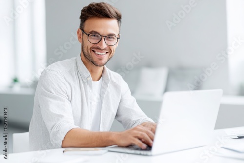 A handsome man in a shirt and glasses is sitting at a white table with a laptop, smiling while working on a digital marketing project in a modern office room with copy space.