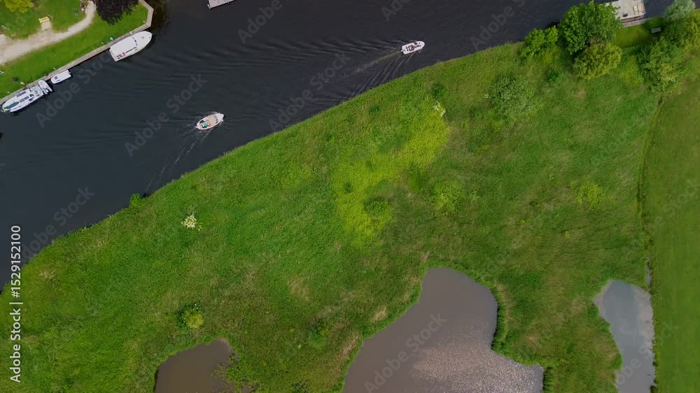 A top-down view of a marina with multiple boats docked along wooden piers, a boat cruising through a narrow waterway surrounded by green parks and natural grassy areas.