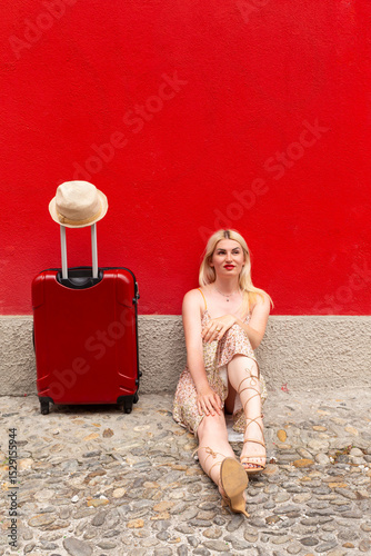 Woman in summer dress travelling with red suitcase in front of a red painted wall