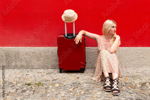 Woman in summer dress travelling with red suitcase in front of a red painted wall