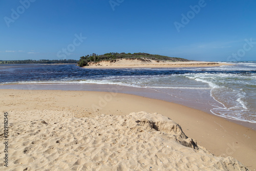 Water flowing between the lake and ocean through a breach in the sandbank at Lake Tyers Beach, Victoria, Australia. Deep blue water, sand dunes and clear sky in a peaceful, sandy seaside landscape.