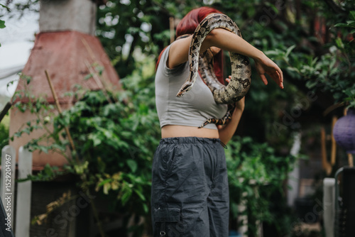 A girl stands in a lush garden, confidently holding a large snake draped over her shoulders. The scene conveys a sense of adventure and connection with nature. photo