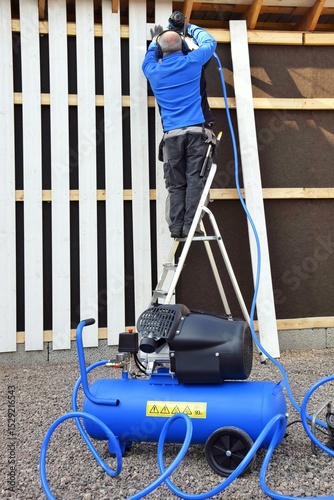 A man stands on a stepladder and holds a pneumatic stapler in his hand - a nail gun for a wooden board, hammering metal nails and staples using a compressor.