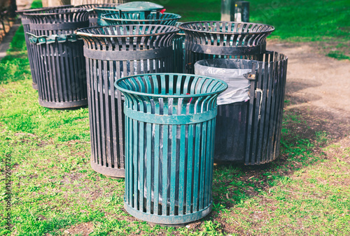 Cylindrical outdoor trash cans with vertical slats, placed on a grassy area. Cans contain plastic liners, suggesting a functional setup in a public park or outdoor space