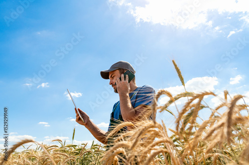 Male agricultural worker multitasking with phone and tablet in cereal field.