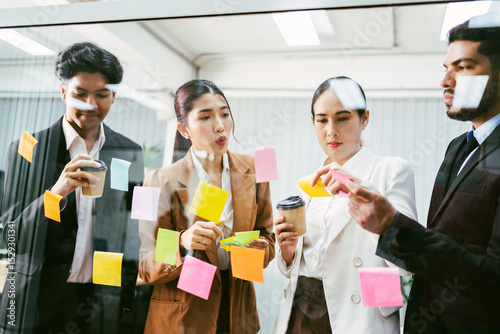 Multicultural office team collaborates in a brainstorming session, placing sticky notes on a glass wall. Business planning, creativity, and teamwork in a modern work environment with coffee in hand.