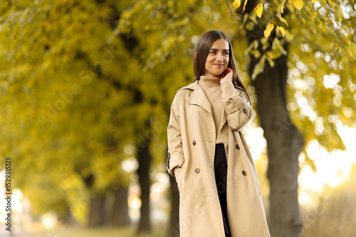 Portrait of pretty young woman in beige trench coat looking at camera relaxing at park during autumn season. Stylish happy natural beauty girl in seasonal casual outfit outdoor, fashion style trend