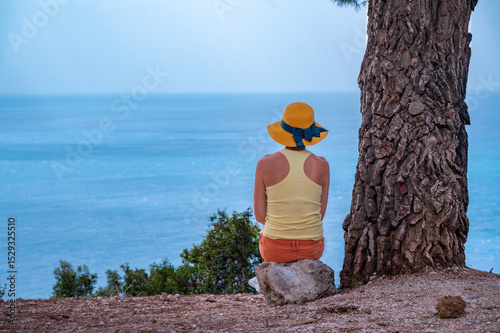Young woman seats next a tree looking towards the sea
