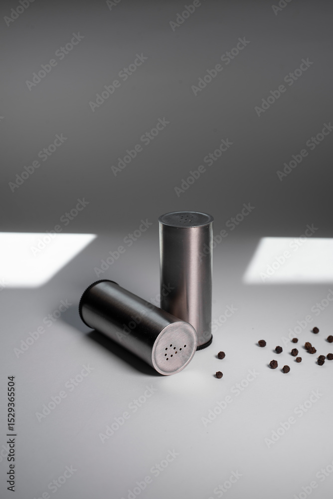 Fototapeta premium Metal pepper shaker and salt shaker, peppercorns next to a white background with light from the sun