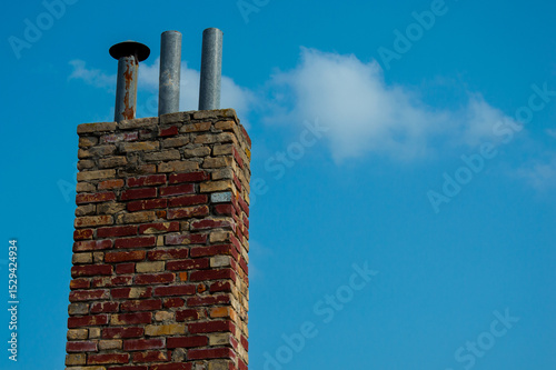 A tall brick chimney with exposed metal flues rises into the clear blue sky on a sunny afternoon, highlighting architectural details